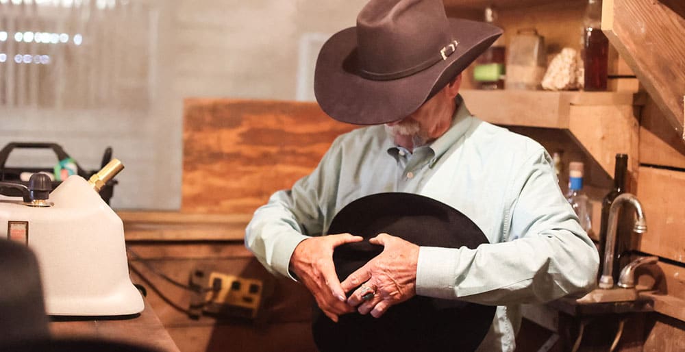 A cowboy wearing a black hat, holding and shaping a black hat.